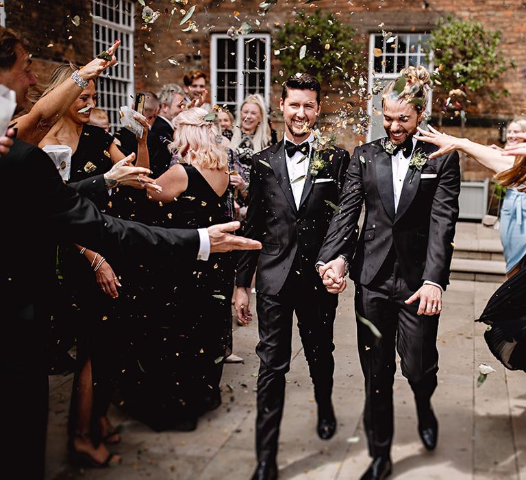 Grooms in black tie walk through colourful confetti outdoors on their wedding day