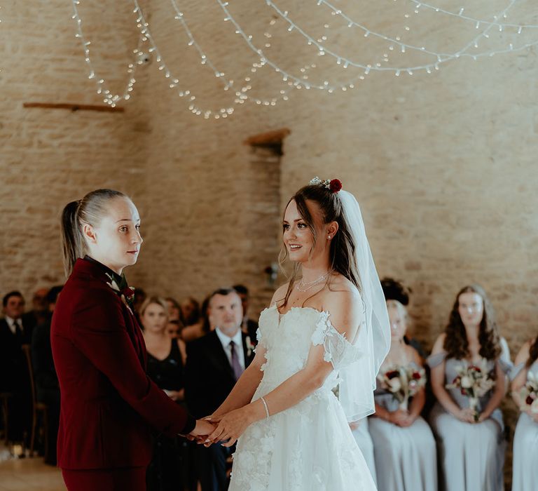 Bride in a burgundy suit with black shirt stands at the altar holding hands with bride in a strapless flower wedding dress and red flower hair accessory