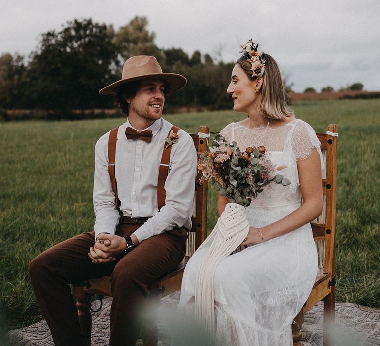 Groom in a striped shirt and brown bow tie, braces and hat sitting on a chair with the bride in a boho wedding dress and dried flower crown 