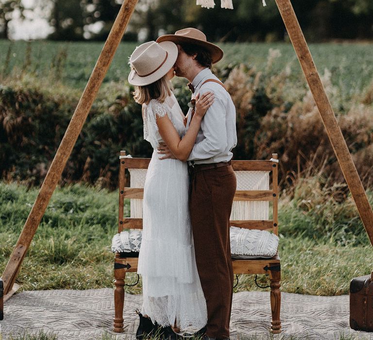 The bride and groom wearing chic and stylish hats for a boho wedding sharing a kiss 