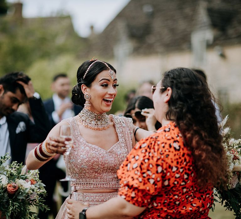 Panjabi Bride in golden bridal Lehnga and Indian bridal jewellery hugs guest while drinking champagne