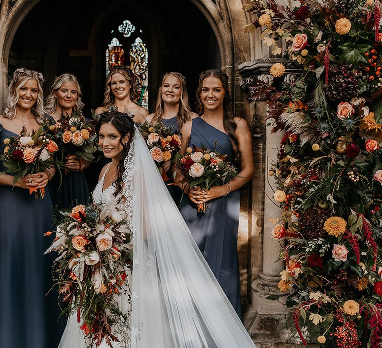 Bridesmaids in navy blue dresses in mismatched styles standing with the bride in between the flower columns all holding boho bouquets with pampas grass