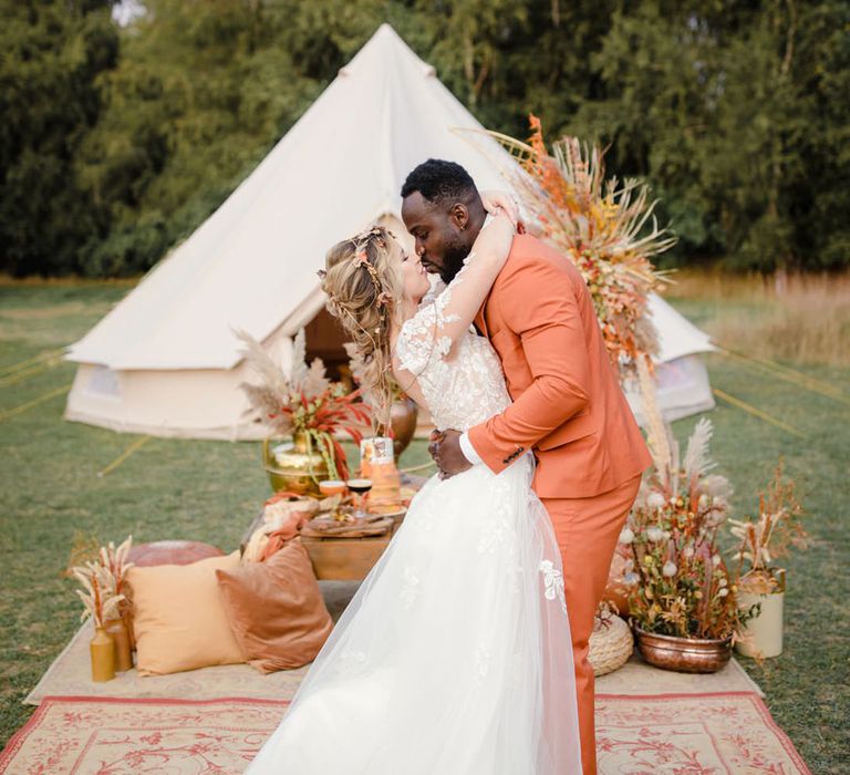 Bride in long sleeved v neck soft ivory dress with lace floral detailing on the bodice and sheer sleeves and groom in burnt orange rust coloured suit with brown tie, pocket square, brown accessories and dried flower, rose and pampas grass boutonniere embracing and kissing on burnt orange rug with autumnal coloured pillows, dried floral arrangements and large white tipi in the background