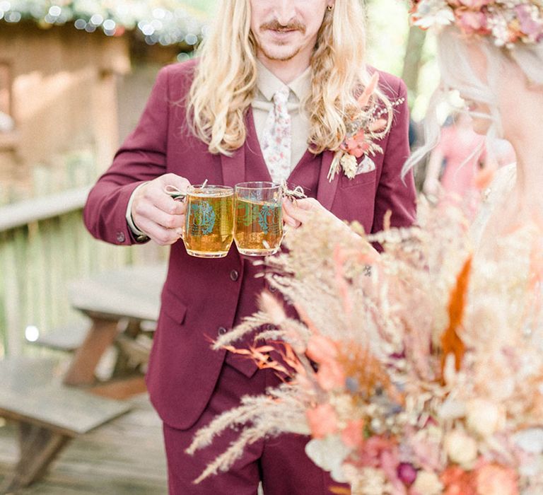 Groom in maroon suit and floral tie holds beer to his bride on their wedding day