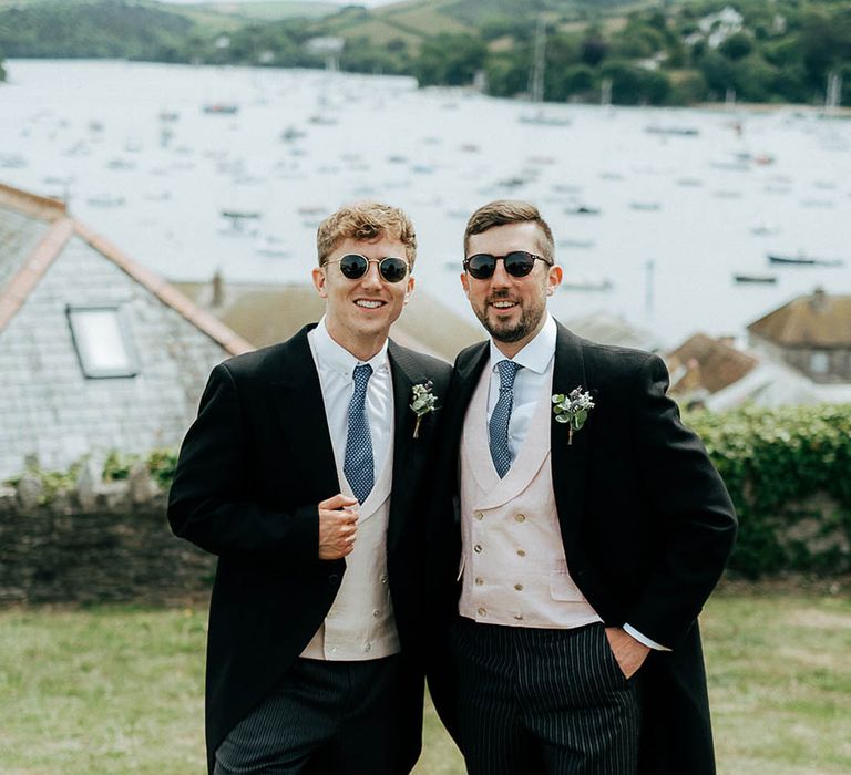 Groom in sunglasses and his groomsmen wear morning suits with blue ties and floral buttonholes 