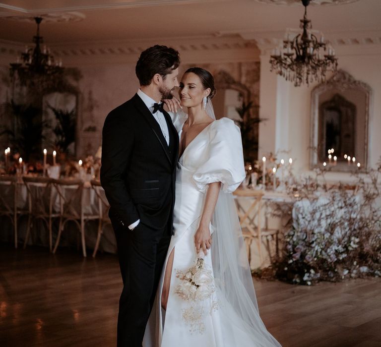 Groom in classic black tux, bowtie and white pocket square embracing bride in v neck puff sleeve wedding dress holding dried flower and white rose bouquet in reception room of Eaves Hall wedding venue  