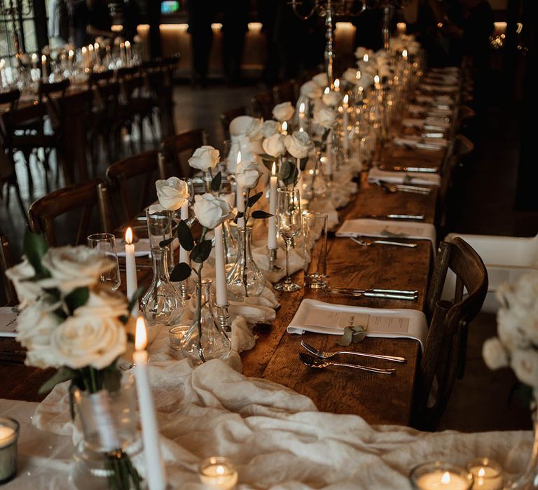 Cosy and luxe wedding tablescape for a rustic wedding with all-white touches including a white table runner, white candles, and white rose arrangements