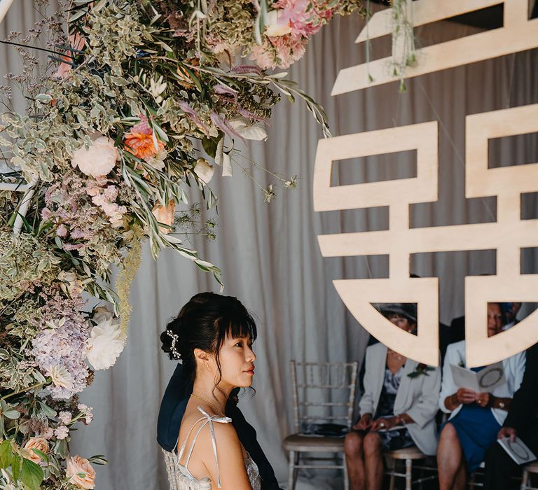 East Asian bride in silver The Vampire's Wife dress sits beside pastel floral arch at Trinity Buoy Wharf