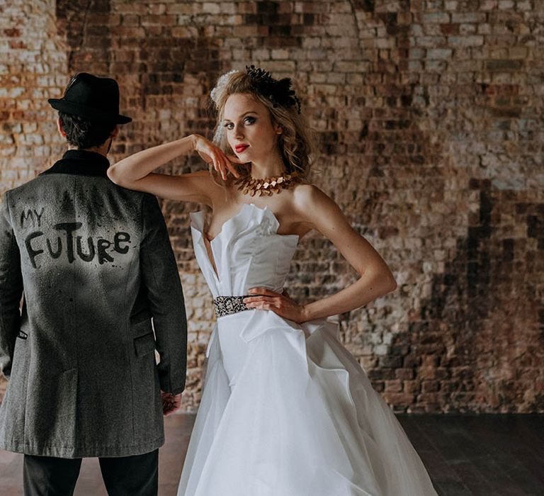 Bride in sculpted bandeau maxi wedding dress, patterned black belt, and large bass necklace standing with groom in personalised dark grey jacket with "my future" graffiti style lettering, black trousers and black fedora hat 