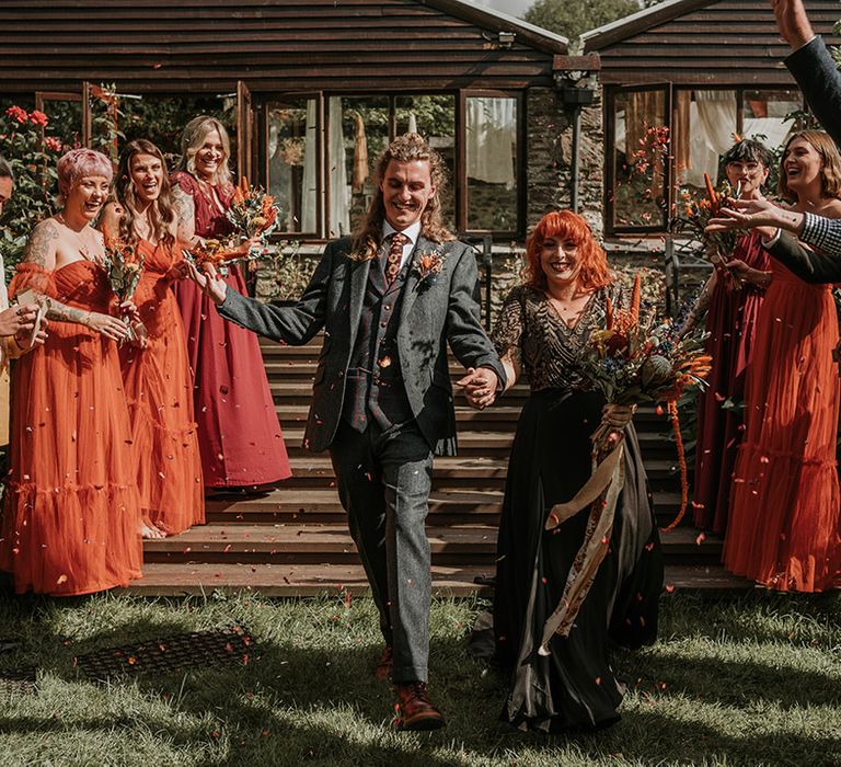 Groom in dark grey three piece suit and burgundy flower patterned tie and bride in embellished black wedding dress holding large orange dried flower bouquet 