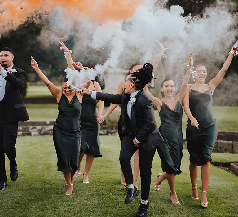 Bride in black snakeskin print tuxedo with black bowtie and white rose boutonniere running with wedding party in strappy satin midi length black bridesmaid dresses and classic black tuxedos with white rose boutonnieres doing smoke bomb wedding photos at Grittleton House wedding venue 