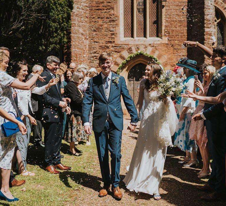 Groom in three piece navy and grey wedding suit having confetti moment with the bride in a traditional lace wedding dress 