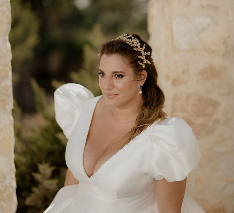 Bride in white satin dress featuring puff-sleeves and a plunge neckline with pearl earrings and a gold leaf bridal crown