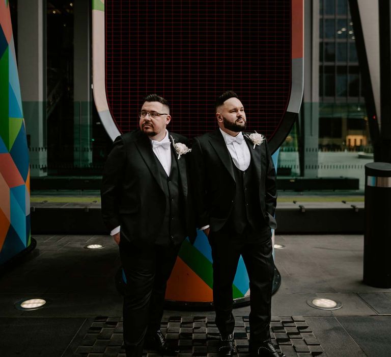 Grooms in complimenting three piece black tuxedos with off white bow ties, white garden rose, lavender twig and dried foliage boutonniere and grooms Louboutins shoes at The Gherkin wedding London 