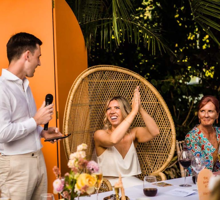 Groom stands up in white shirt and cream trousers reading out his wedding speech with the bride in satin two piece on peacock chair 