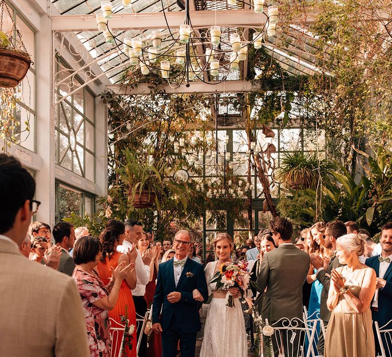 Bride in modern two piece carrying a colourful bouquet with sunflowers walking down the aisle with her father 