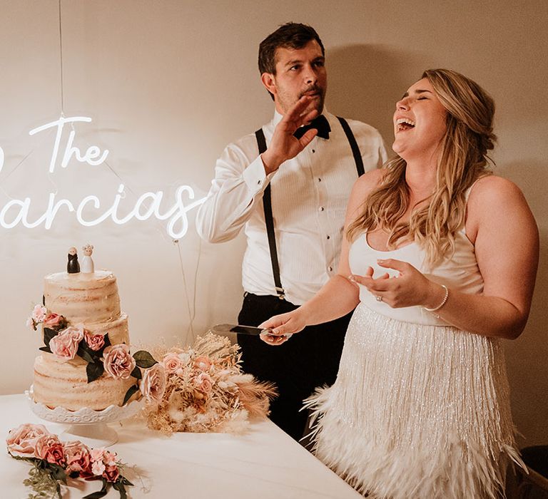 Personalised white neon wedding sign behind rustic semi-naked wedding cake with pink flower decor being cut by the bride and groom 