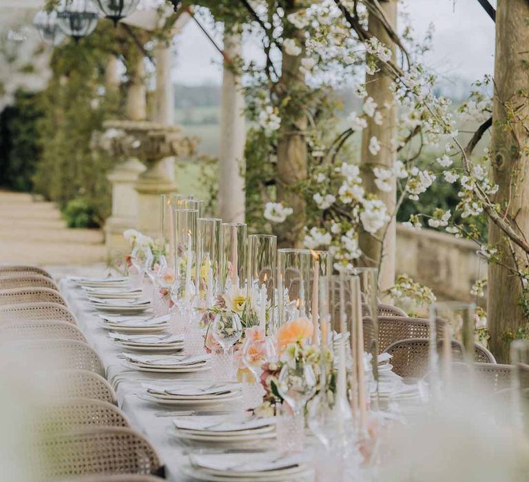 Outdoor al fresco wedding tablescape with blush pink tapered candles in glass candlestick covers on spring inspired wedding tablescape with white ruffled wedding table cloth, white peony, pink garden rose, foliage and blush florals wedding table centrepieces and simple crockery at Euridge Manor wedding