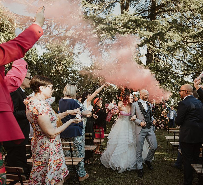 bride in strapless wedding dress and groom in a grey suit walking up the aisle as husband and wife at their outdoor italian villa wedding ceremony with pink smoke flares