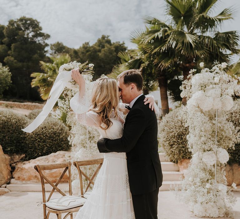 bride in a long bell sleeve lace wedding dress waving her white wedding bouquet tied with ribbon in the air as she kisses her groom in a tuxedo at pure house ibiza wedding