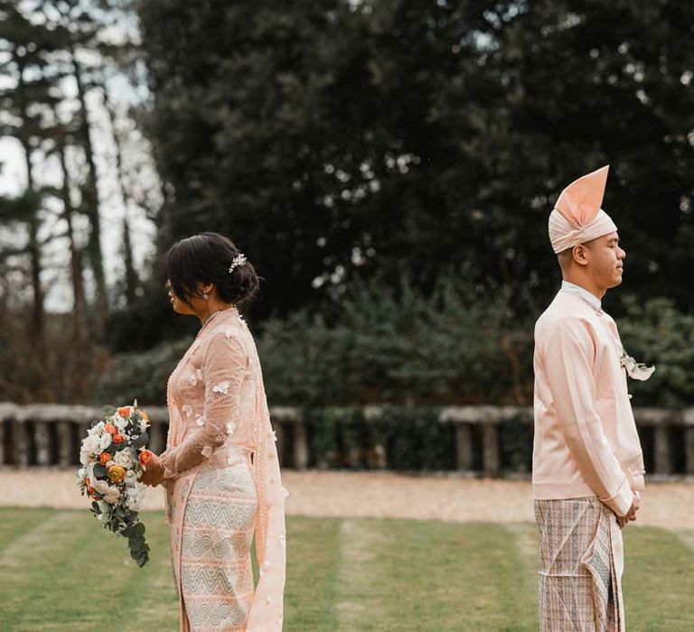 Groom in baby pink htaingmathein and bride in floral 3D applique pink and rose gold htamane back to back doing first look wedding photos at Burmese wedding