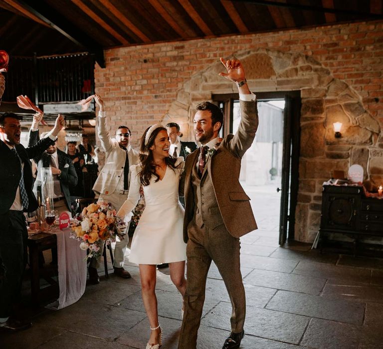 Bride in long sleeve short wedding dress, pearl bridal headband and platform pearl wedding heels walking into reception room of Willow Marsh Farm Loughborough with groom in three piece chocolate brown grooms suit 