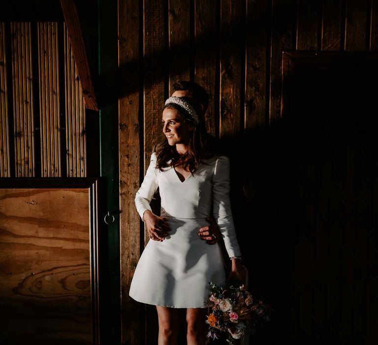 Bride standing in the shadows in long sleeve short wedding dress, pearl bridal headband and platform pearl wedding heels with groom behind her