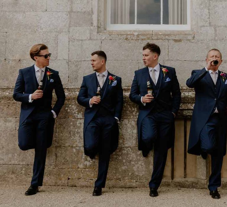 Groom and groomsmen in navy groom morning suits with grey ties and blush rose boutonnieres holding beers at Elmore Court Gloucestershire 