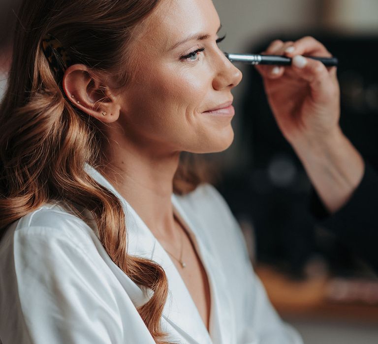 Bride in white robe sits getting ready for the day getting makeup done 