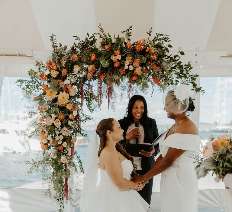 Black bride in an off the shoulder wedding dress and blush veil holding hands with her bride in a strapless wedding dress during their same sex wedding ceremony 