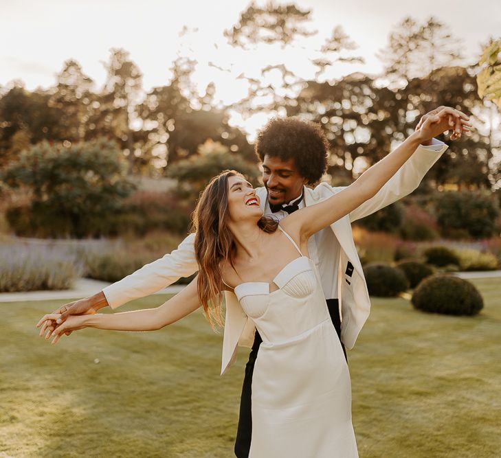 Black groom with afro hair and white tuxedo jacket holding hands with his bride with long brown hair and red lipstick in a satin dress with cup detail and thin straps 