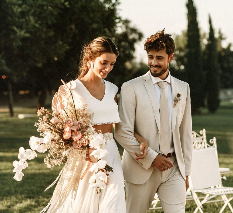 Boho bride in separates holding an orchid bouquet walking down the aisle with her groom in a beige suit 