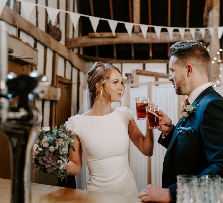 Groom in a navy suit and bride in a fitted wedding dress with appliqué shoulder detail drinking Mulled Wine 