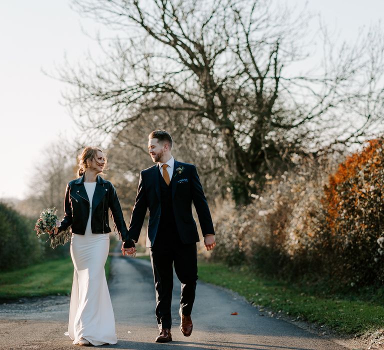 Bride and groom walking down a country lane in a navy suit and orange tie and fitted wedding dress and leather jacket 