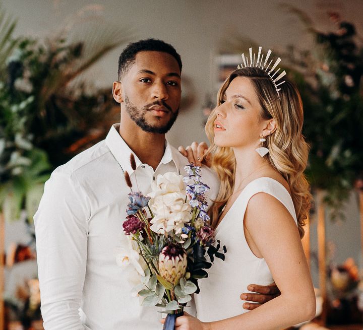 Groom in leather trousers and white shirt and bride in a fitted dress wearing a gold crown and holding a purple wedding bouquet 