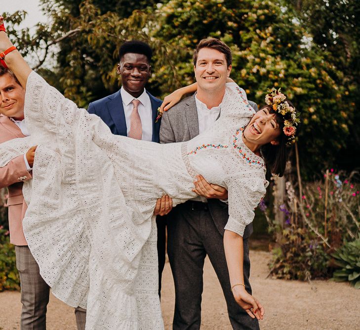 Groom and groomsmen picking up the bride in a white dress with colourful embroidery, flower crown and red shoes 