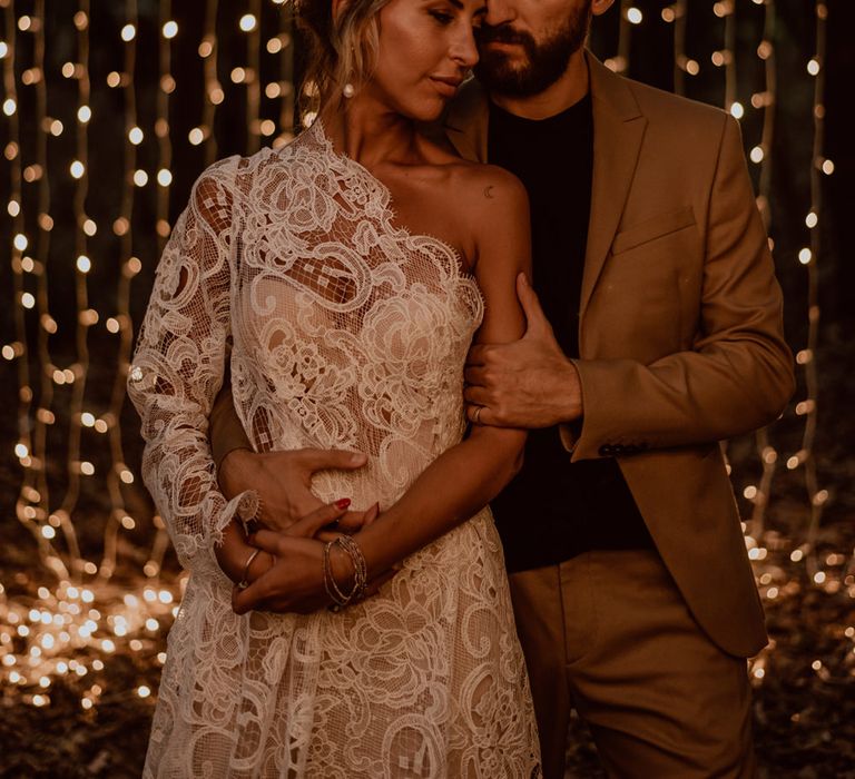 Bride in a one shoulder wedding dress standing in front of a fairy light curtain with her groom in a brown suit