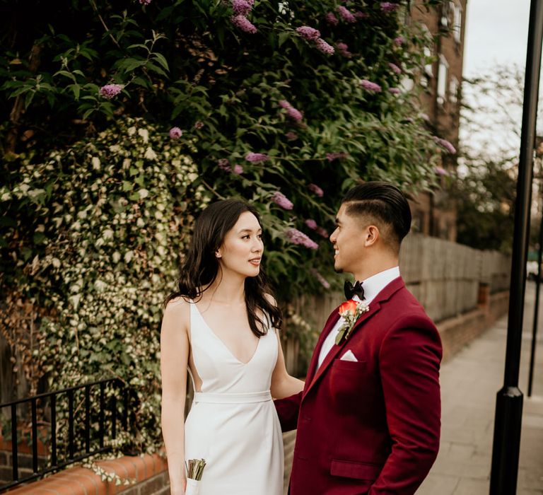 Bride in a Sarah Seven wedding dress holding a red and white rose bouquet with her husband in a burgundy jacket and bow tie 