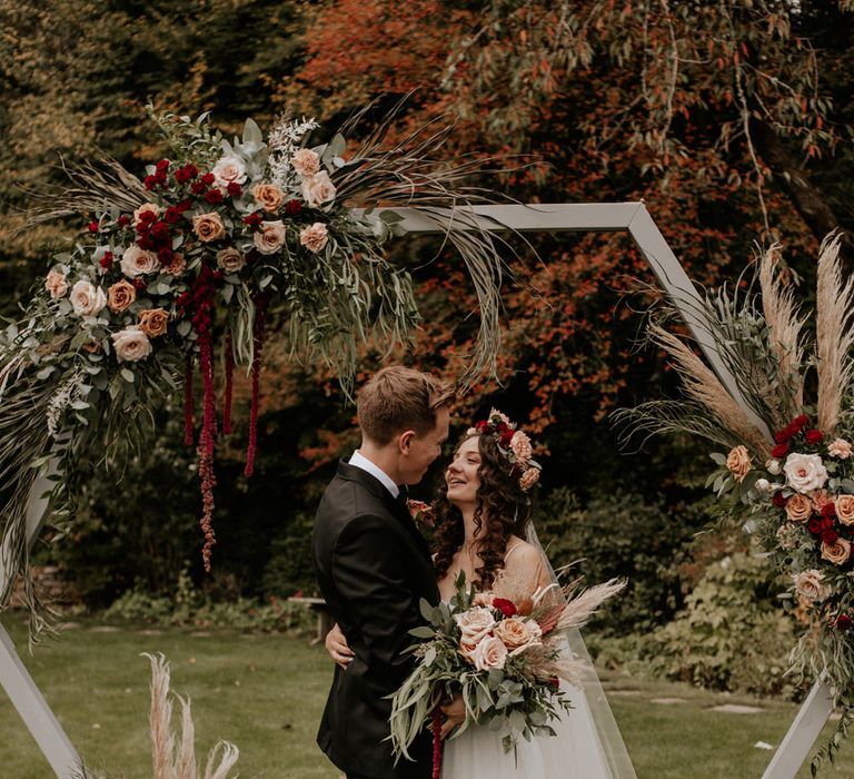 Groom in a black tuxedo looking at his boho bride in an tulle skirt wedding dress with flower crown and veil in front of a wooden frame decorated with flowers 