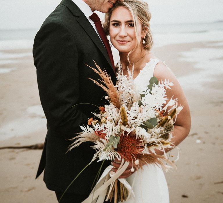 Bride & groom embrace as they stand together on the beach