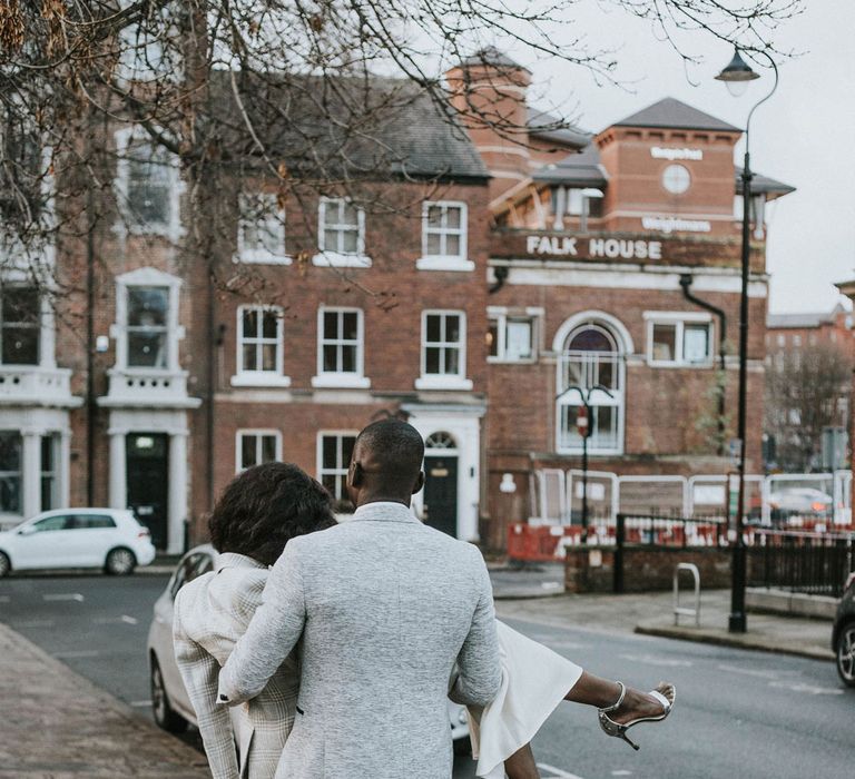 Groom in grey suit jacket, black trousers and brown shoes carries bride in white dress, grey checked blazer and silver heeled sandals down a street in Leeds