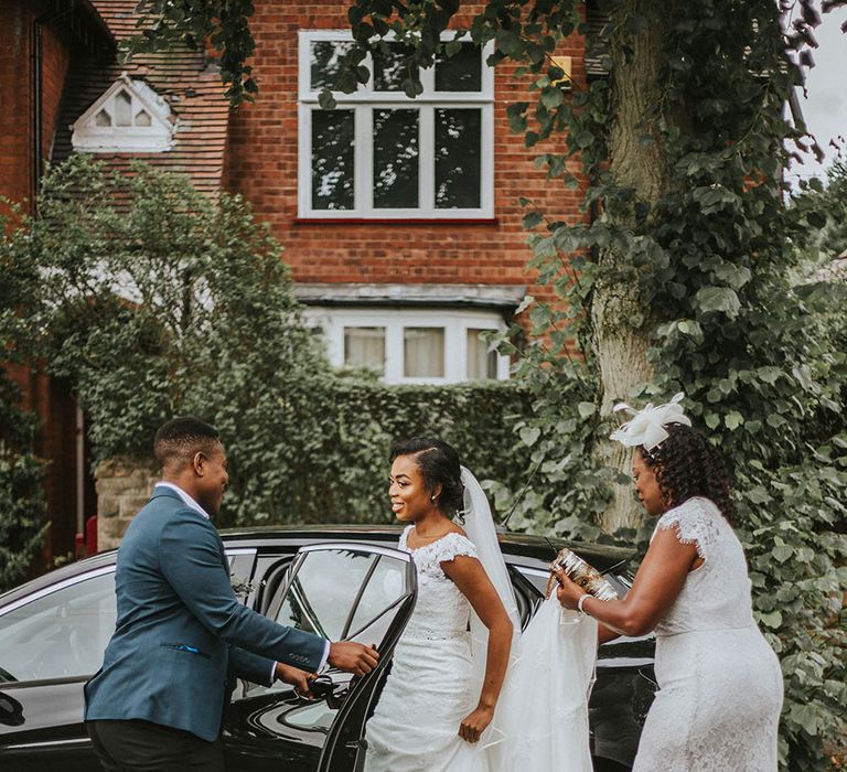 Mother of the bride in a white lace midi dress and fascinator holding her daughters train as she gets into the wedding car 