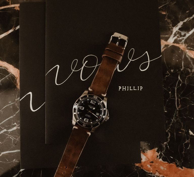 Men's watch with a brown strap, laid on top of black wedding vows booklets with calligraphy font