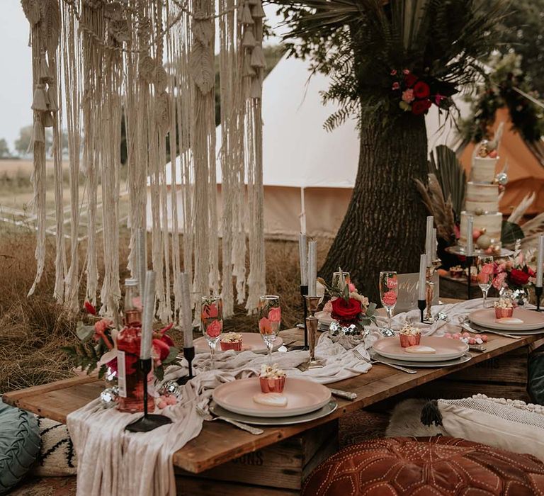 Low wooden wedding tablescape with beige table runner, long silver candles, pink pastel colours and macrame hanging outside at festival themed wedding