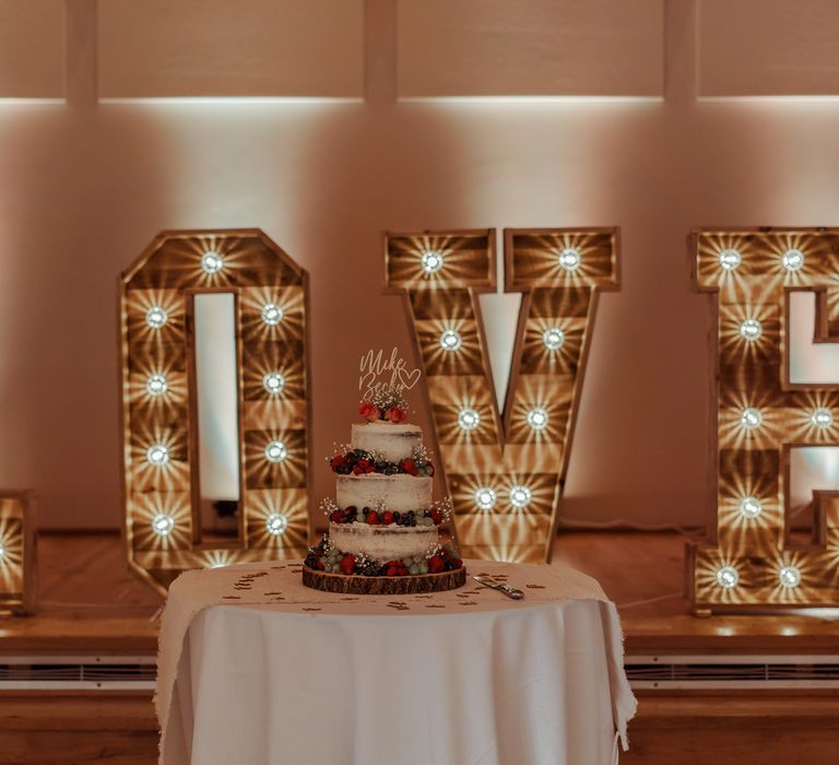 Lit up LOVE wedding sign sits behind naked wedding cake with fruit decor