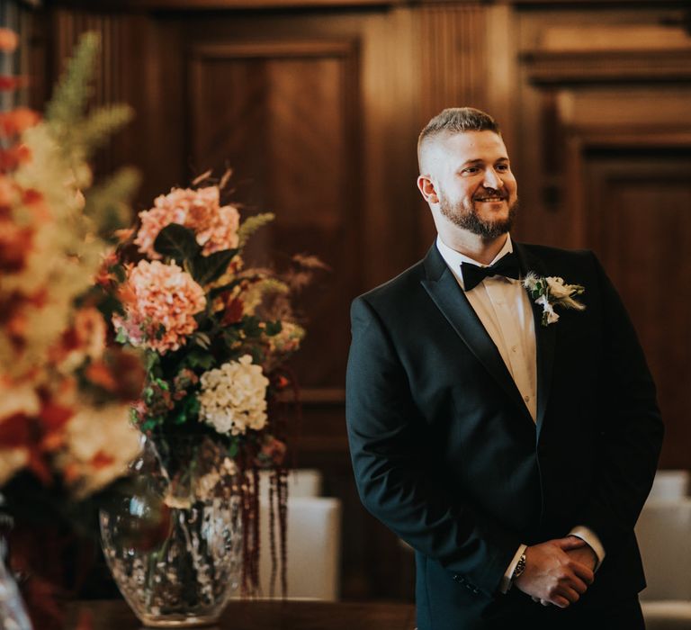 Groom awaits bride and smiles as he looks on whilst stood by large and colourful floral arrangements 