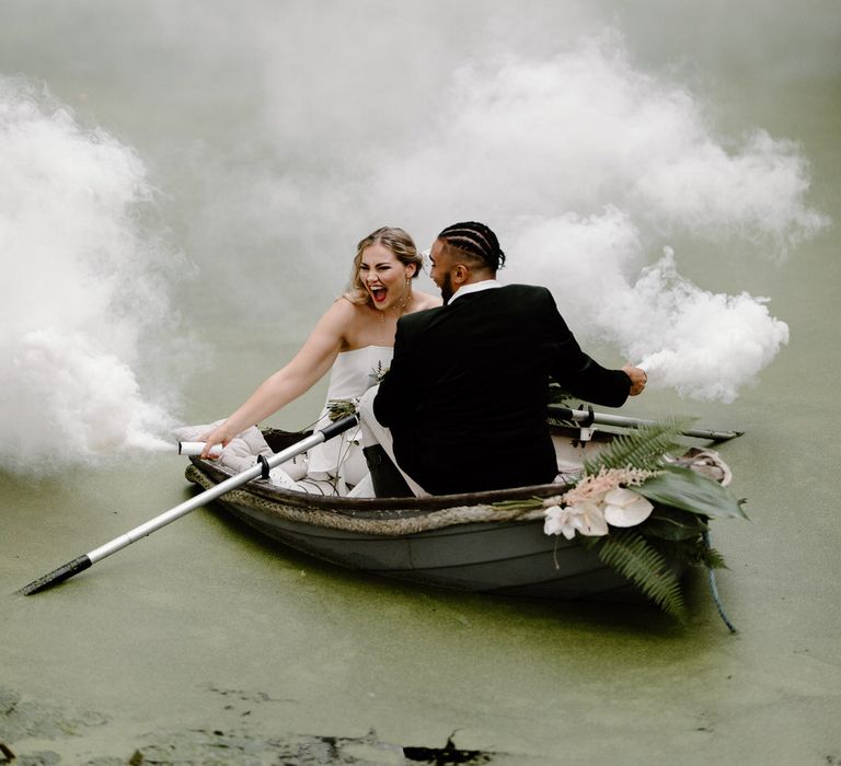 Bride and groom on a green algae covered lake in a row bow, holding white smoke flares