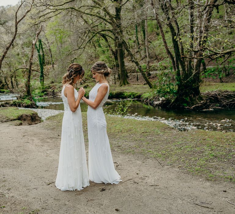 Brides claps hands and stand facing one another whilst stood outdoors surrounded by woodland