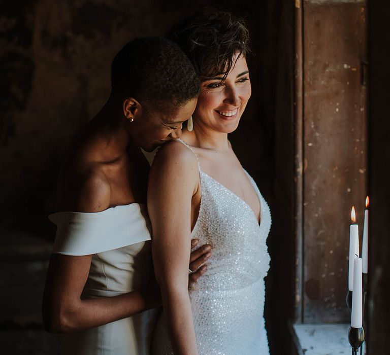 Black bride in a strapless wedding dress kissing her brides shoulder in a sparkly wedding dress as she looks out the window 