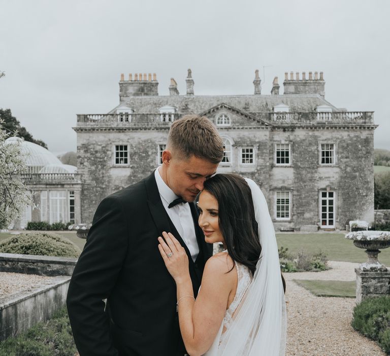 Bride in white Pronovias wedding dress and veil holds hand on grooms chest as they stand in front of Came House Dorset after their wedding